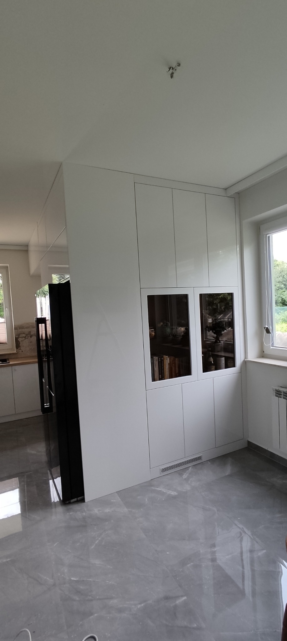 Biała zabudowa meblowa z przeszklonymi witrynami, wbudowana lodówka obok okna z grzejnikiem, szara marmurowa podłoga.