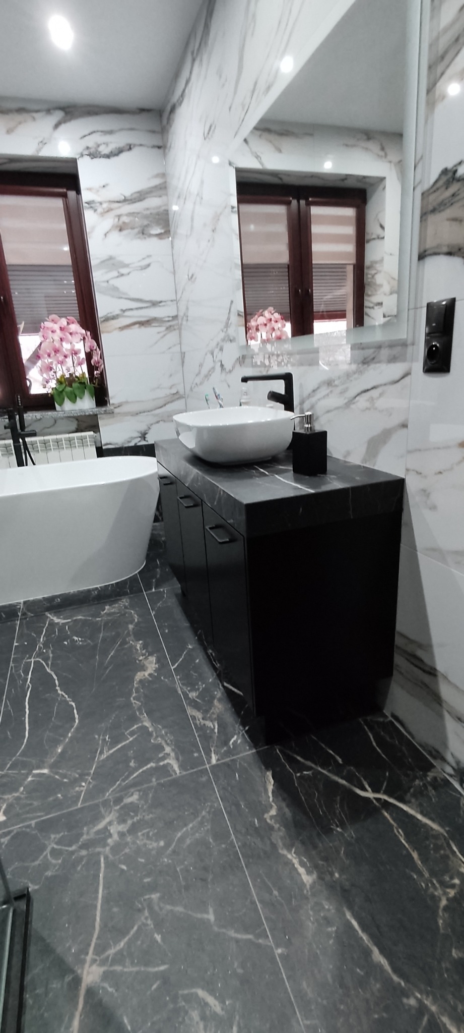 Nowoczesna łazienka z czarną szafką pod umywalką nablatową, marmurowymi płytkami na ścianach i podłodze oraz wolnostojącą wanną.