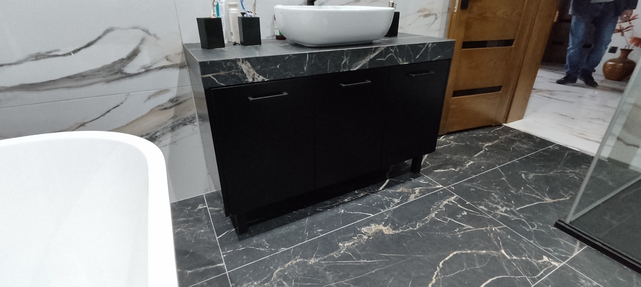 Czarna szafka łazienkowa z marmurowym blatem, umywalka nablatowa i akcesoria, na tle marmurowych płytek ściennych i podłogowych.