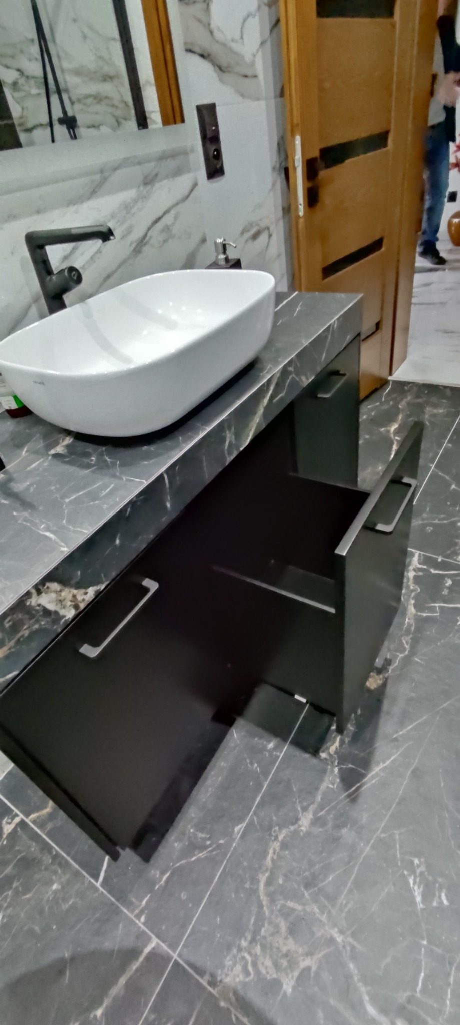 Łazienka z umywalką nablatową i czarną szafką z otwartą szufladą na tle marmurowej ściany i podłogi.