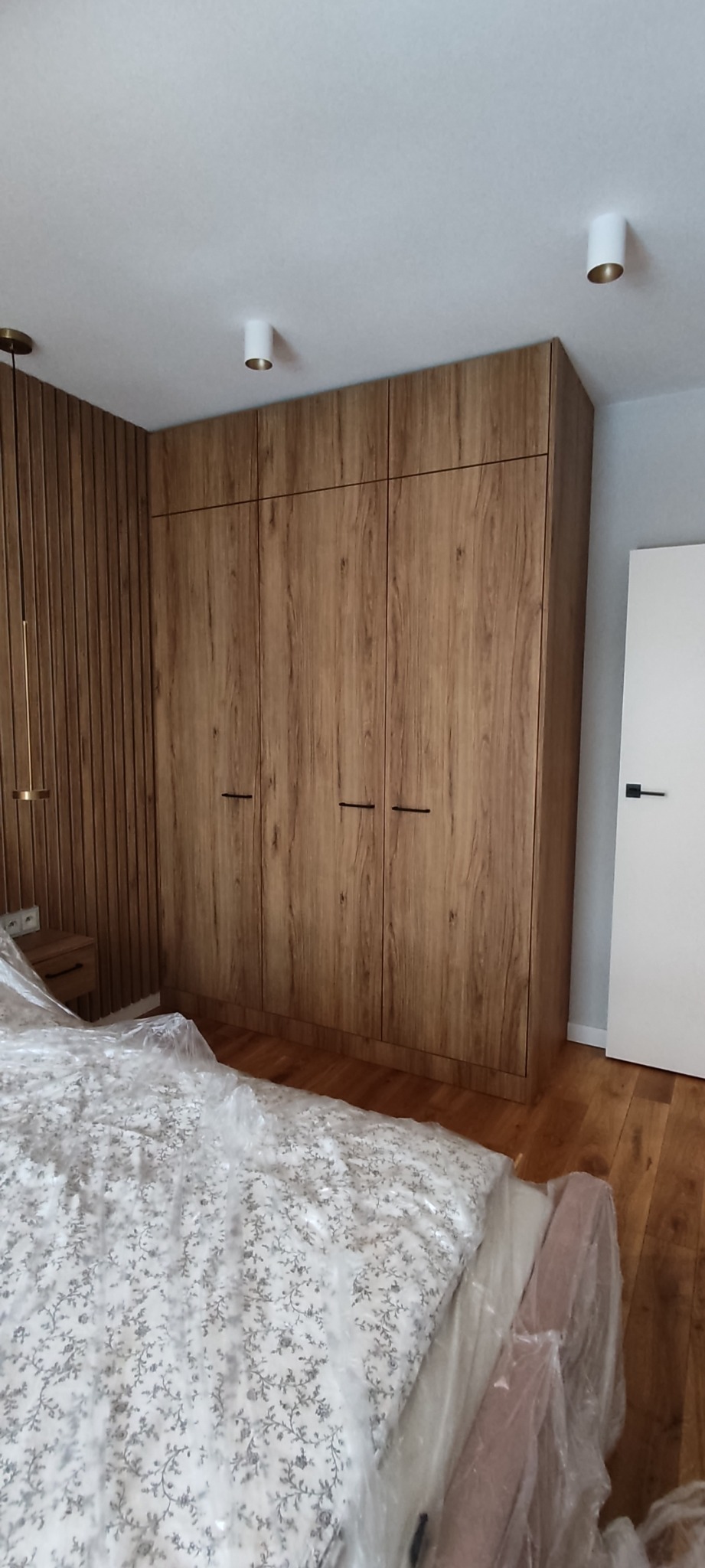 Wysoka szafa z frontami imitującymi drewno, z czarnymi uchwytami, dopasowana do ściany z drewnianymi panelami w sypialni, łóżko przykryte folią ochronną.