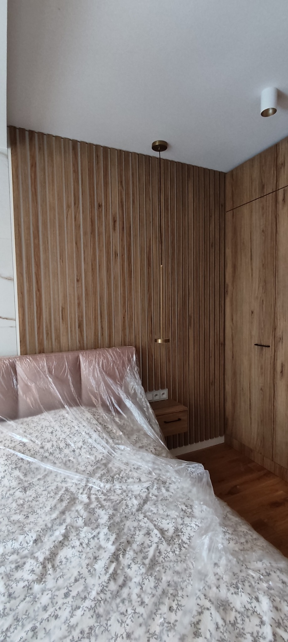 Sypialnia z drewnianą boazerią ścienną, szafą w zabudowie i łóżkiem przykrytym folią ochronną.