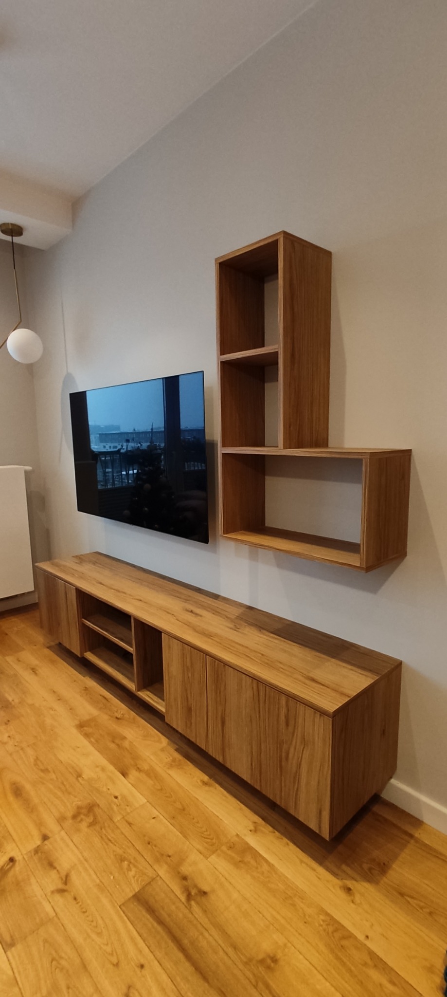 Drewniana zabudowa ściany z szafką RTV zamykaną na fronty i wiszącymi półkami, oświetlenie sufitowe w formie kuli na złotym ramieniu, parkiet w jodełkę.