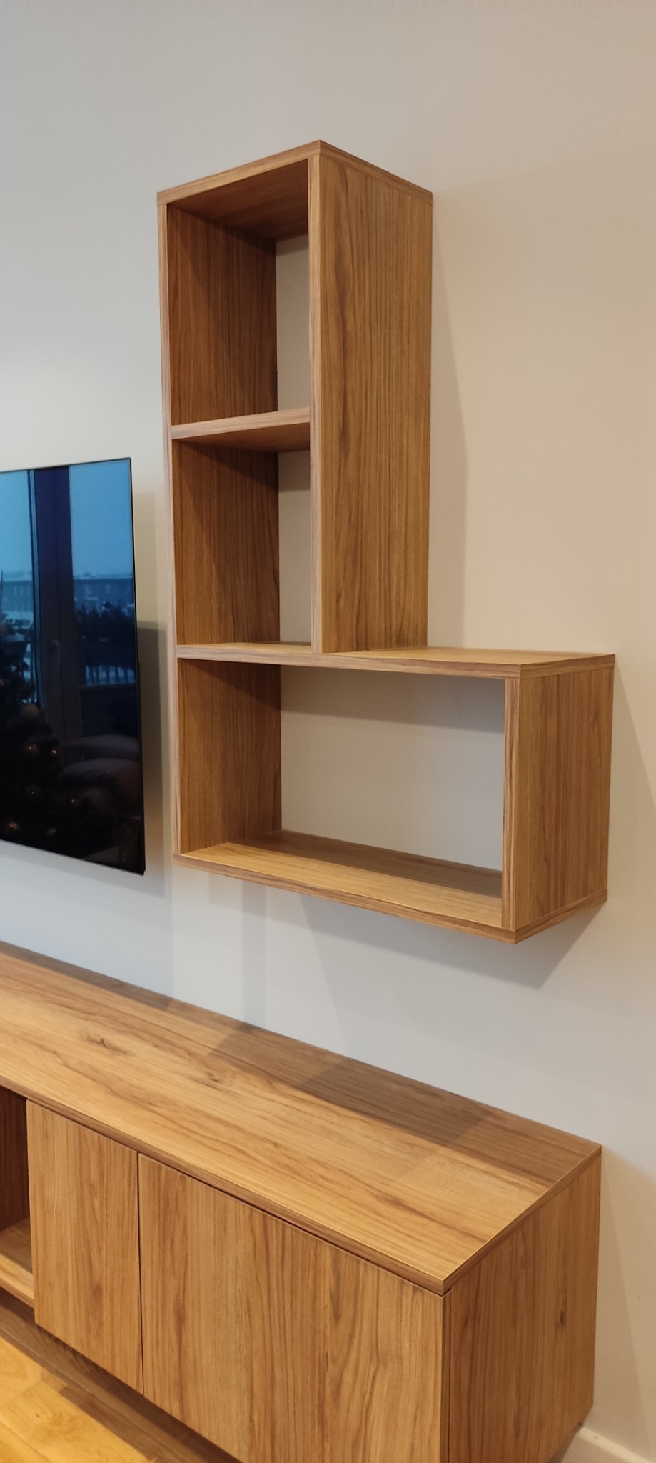 Drewniana szafka RTV z wiszącymi półkami w kształcie kostek, widok z boku na tle ściany i telewizora.