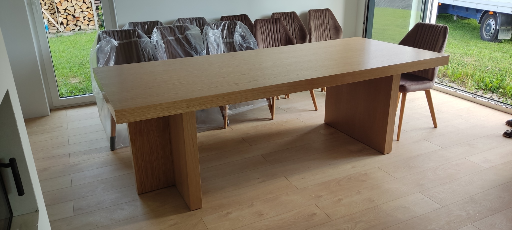 Jasny, prostokątny stół z litego drewna z grubym blatem i nogami, ustawiony w nowoczesnym wnętrzu na podłodze z paneli, z krzesłami obitymi brązowym welurem, niektóre jeszcze w folii ochronnej...