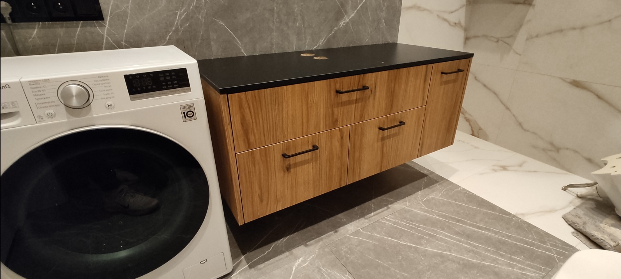 Szafka łazienkowa na wymiar z drewnianymi frontami i czarnym blatem, obok pralka, szare płytki podłogowe z marmurkowym wzorem.