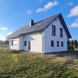 Nowoczesny, parterowy dom z białym tynkiem i ciemną dachówką, widoczny z perspektywy trawnika w słoneczny dzień.