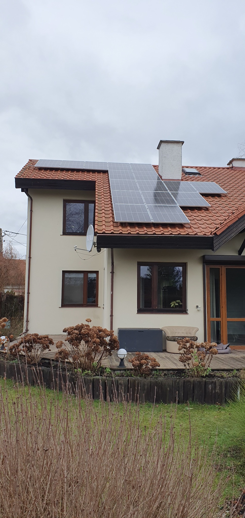 Dwukondygnacyjny dom murowany z czerwonym dachem, na którym zamontowano panele fotowoltaiczne; widoczna antena satelitarna, drewniany taras z meblami ogrodowymi i uschniętymi kwiatami.