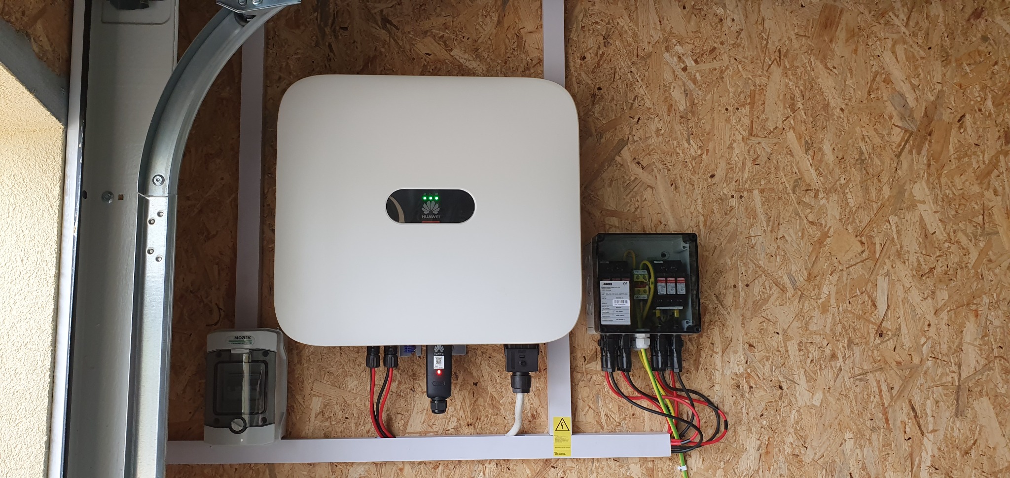 Biały inwerter solarny Huawei z podłączonymi kablami i zabezpieczeniami, zamontowany na ścianie z płyty OSB w Gdyni. Widoczne zielone diody sygnalizujące pracę urządzenia i skrzynka z bezpiecznikami.