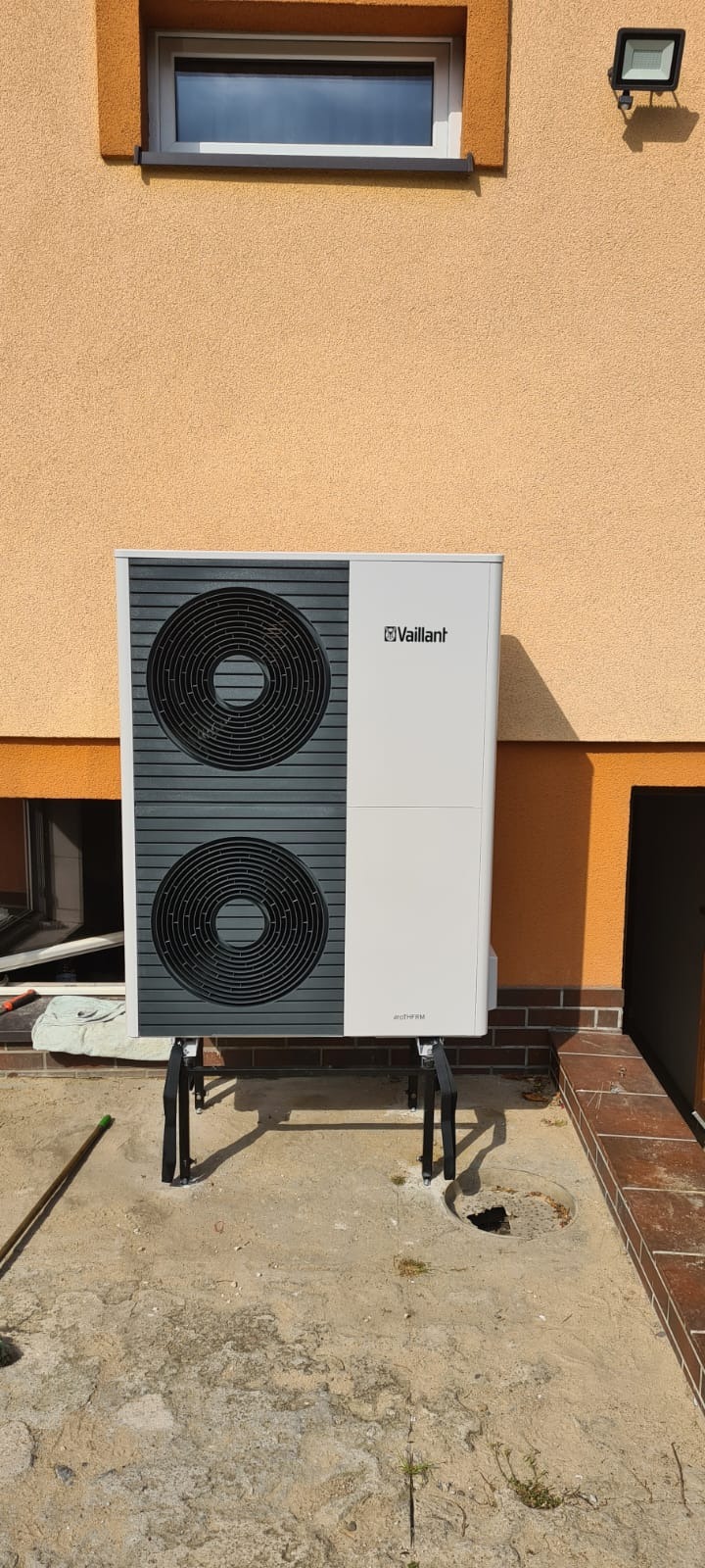 Biała pompa ciepła Vaillant zamontowana na czarnych wspornikach przy beżowej ścianie budynku z widocznym oknem i lampą zewnętrzną.