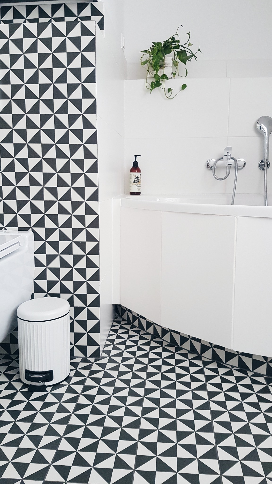 Minimalistyczna łazienka z białą zabudową wanny, wzorzystymi czarno-białymi płytkami na podłodze i ścianie, oraz rośliną zwisającą z sufitu w doniczce.
