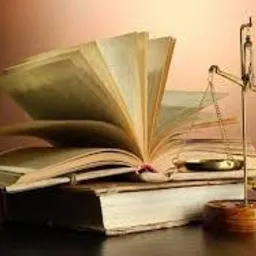 Otwarta, stara książka prawnicza leżąca na stosie innych ksiąg, obok złote wagi sprawiedliwości na drewnianym biurku, oświetlona ciepłym światłem.