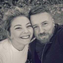 Linia Natury - Portret uśmiechniętej kobiety z wysoko upiętymi włosami i brodatego mężczyzny w bluzie, zrobiony w czerni i bieli, z naturalnym tłem.