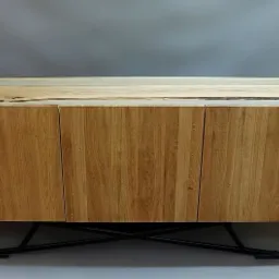 Nowoczesna szafka RTV z jasnego drewna z żywicą epoksydową na blacie, wsparta na czarnych, metalowych nogach w kształcie litery X, na tle szarej ściany.