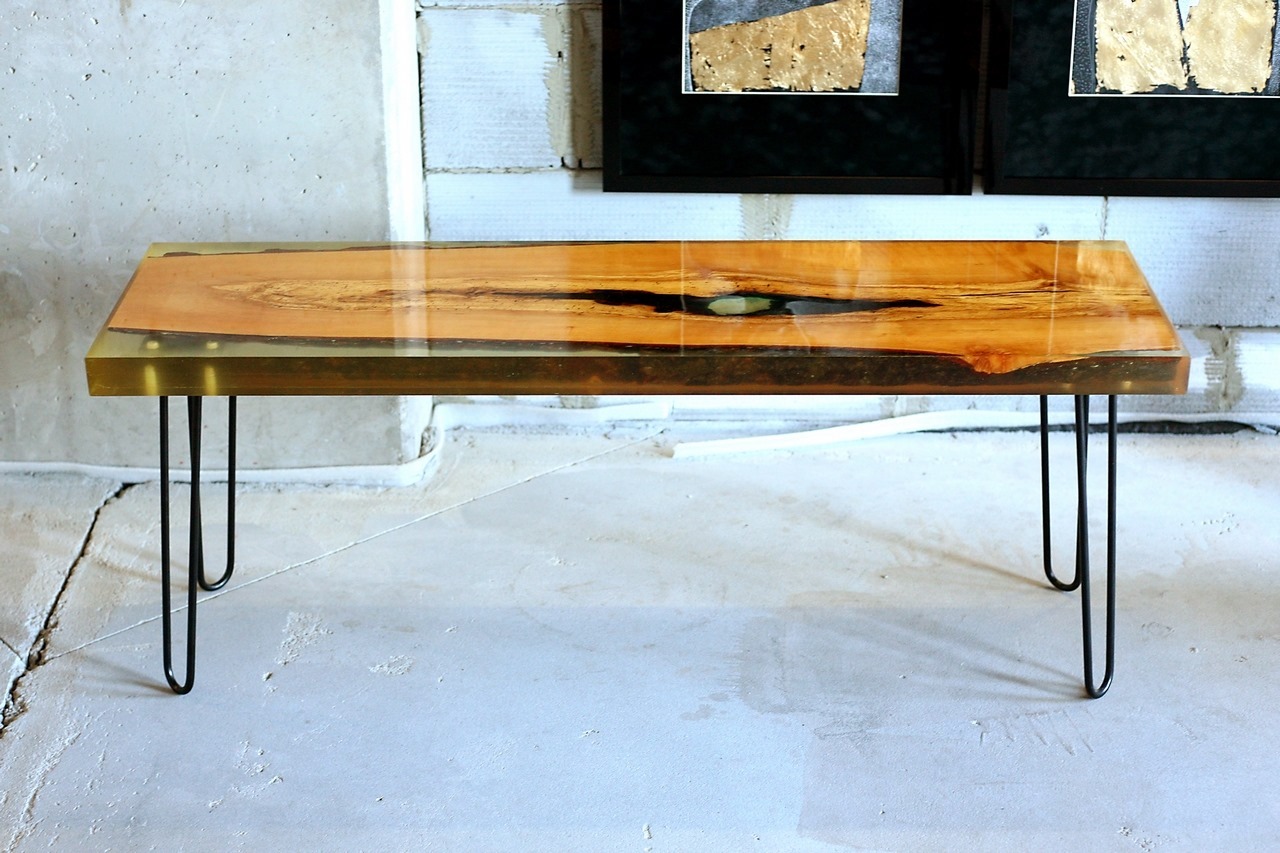 Stolik kawowy z blatem z drewna zatopionego w żywicy epoksydowej, na metalowych nóżkach typu hairpin, na tle surowej ściany z cegły i obrazów.