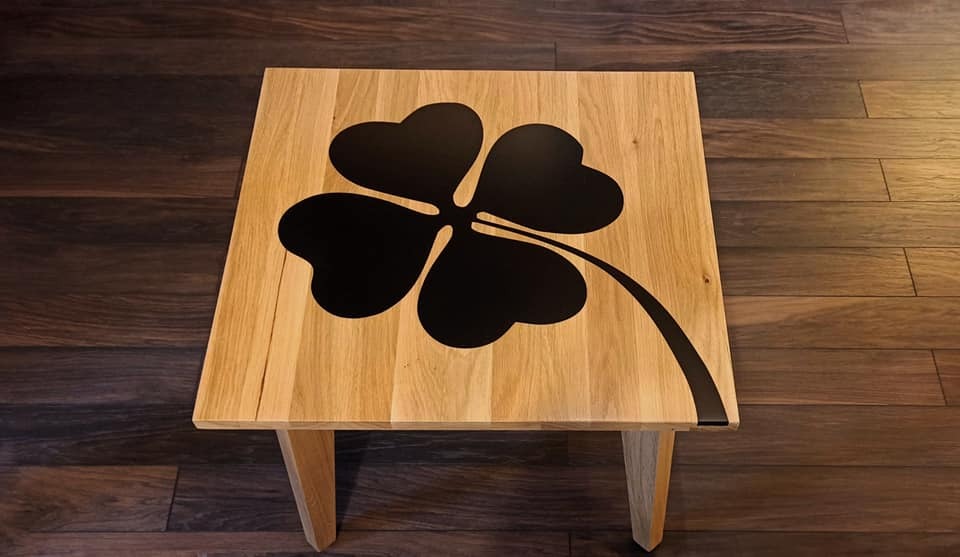 Drewniany stolik kawowy z blatem zdobionym czarną grafiką czterolistnej koniczyny, ustawiony na drewnianej podłodze.