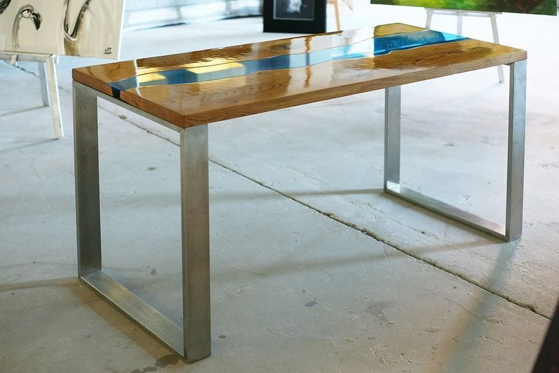 Biurko z blatem z drewna dębowego i żywicy epoksydowej w kolorze niebieskim, na metalowych nogach o kwadratowym przekroju, w tle obrazy na sztalugach.