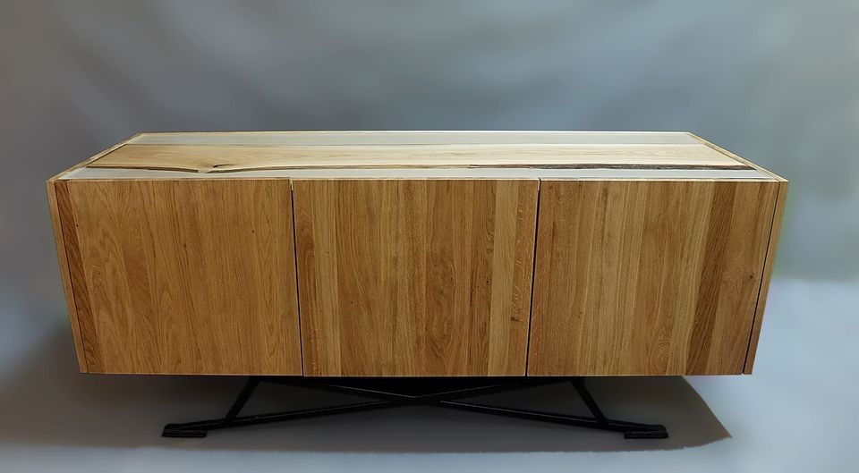 Nowoczesna szafka RTV z jasnego drewna z żywicą epoksydową na blacie, wsparta na czarnych, metalowych nogach w kształcie litery X, na tle szarej ściany.