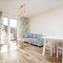 Jasne wnętrze pokoju dziennego z podłogą z paneli, błękitną sofą z poduszkami, białym stołem i krzesłami, oraz zasłonami w kwiatowy wzór przy oknie balkonowym, oświetlone wiszącymi lampami...