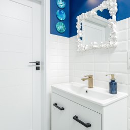 WMK odbiory i remonty mieszkań oraz domów - Nowoczesna łazienka z białą szafką pod umywalką, złotą baterią i dekoracyjnym lustrem w białej ramie na tle ściany z niebieskim akcentem i białymi płytkami.