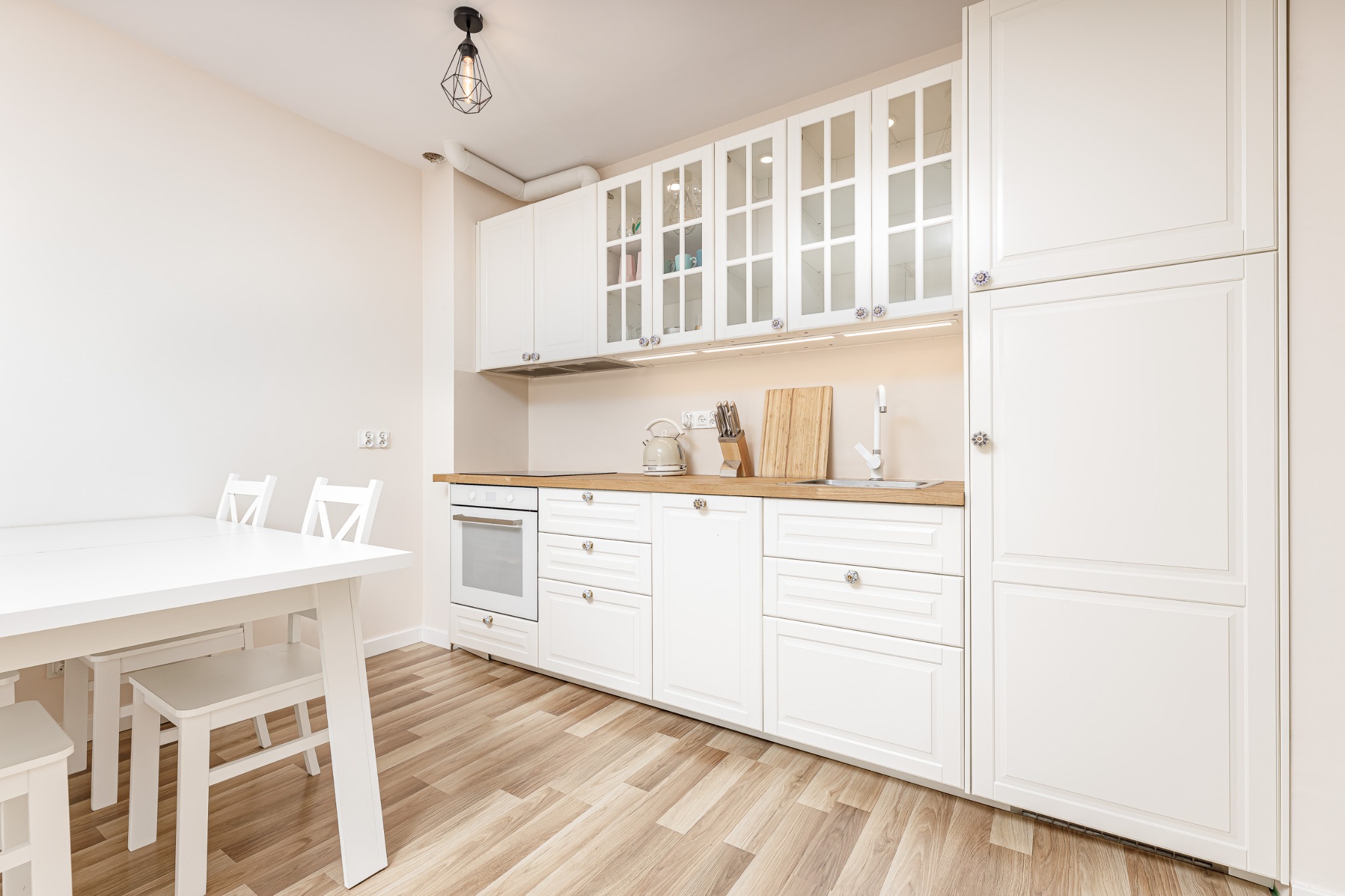 Jasna kuchnia z białymi meblami, drewnianym blatem, białym stołem z krzesłami i lampą w stylu industrialnym.