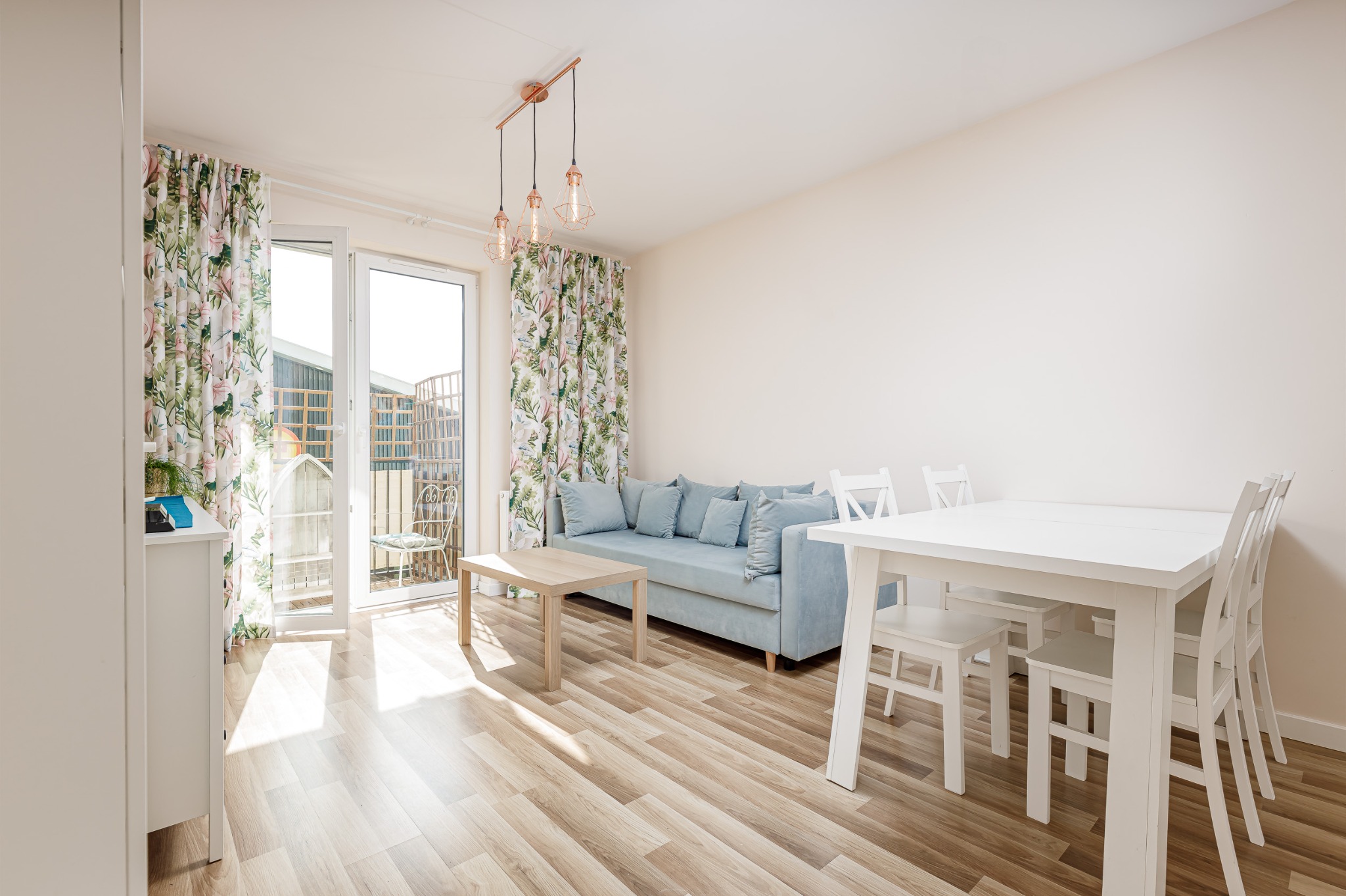 Jasne wnętrze pokoju dziennego z podłogą z paneli, błękitną sofą z poduszkami, białym stołem i krzesłami, oraz zasłonami w kwiatowy wzór przy oknie balkonowym, oświetlone wiszącymi lampami...