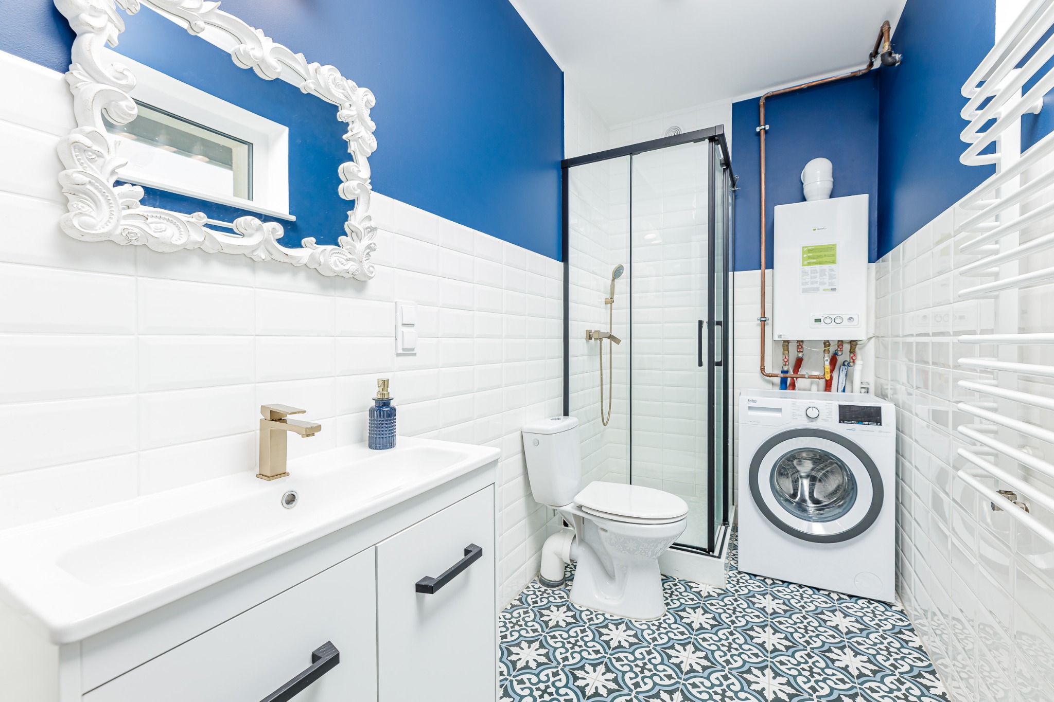 Niebiesko-biała łazienka z kabiną prysznicową, umywalką z szafką, toaletą, pralką i dekoracyjną ramą lustra.