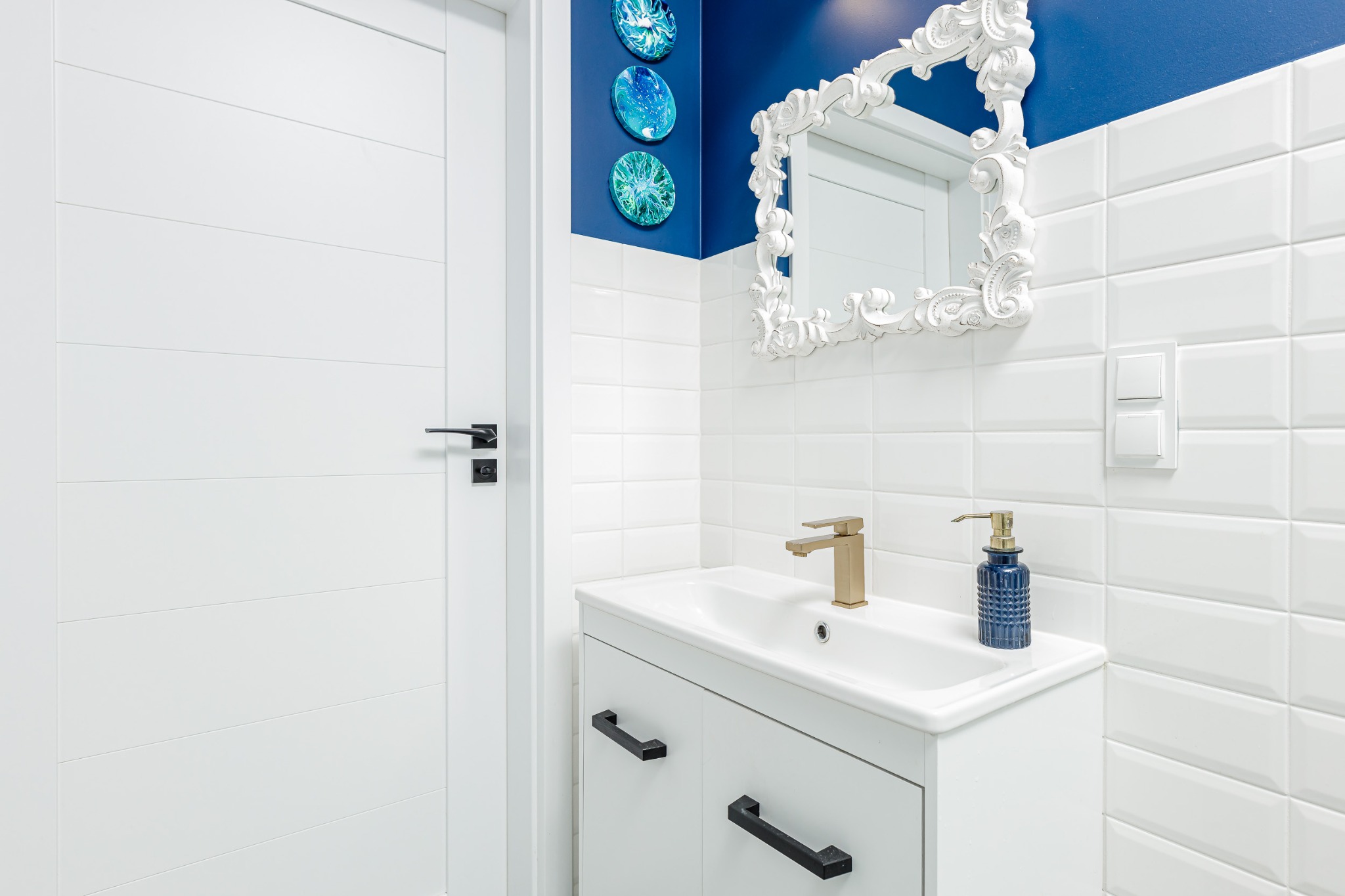Nowoczesna łazienka z białą szafką pod umywalką, złotą baterią i dekoracyjnym lustrem w białej ramie na tle ściany z niebieskim akcentem i białymi płytkami.