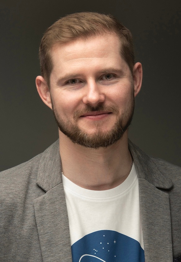 Portret uśmiechniętego mężczyzny w szarej marynarce i białej koszulce z nadrukiem, na szarym tle.