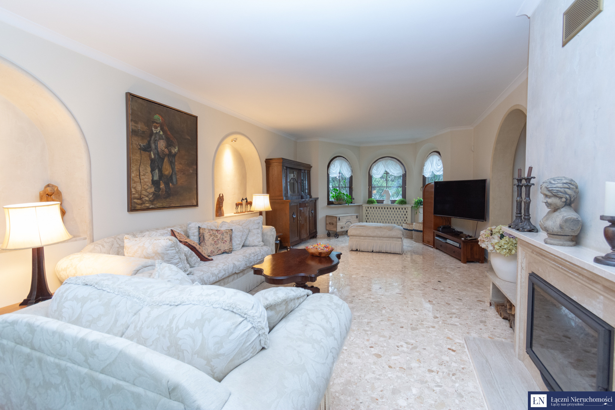 Przestronny salon z jasnym, wzorzystym dywanem, kominkiem i antycznym kredensem, oświetlony lampą stojącą i dodatkowo doświetlony przez okna z dekoracyjnymi zasłonami.