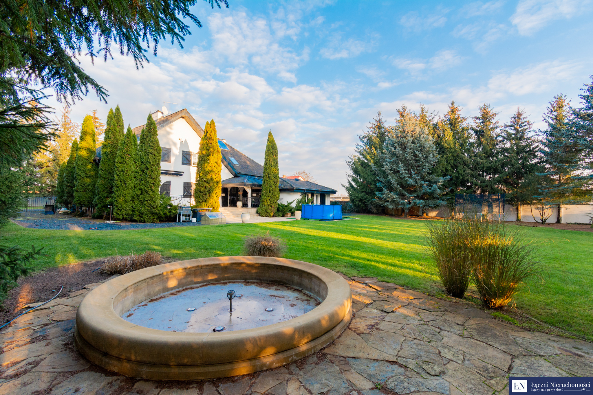 Rozległa posiadłość z białym domem, zielonym trawnikiem, fontanną bez wody na pierwszym planie, otoczona tujami i drzewami iglastymi, widok z kamiennego tarasu.