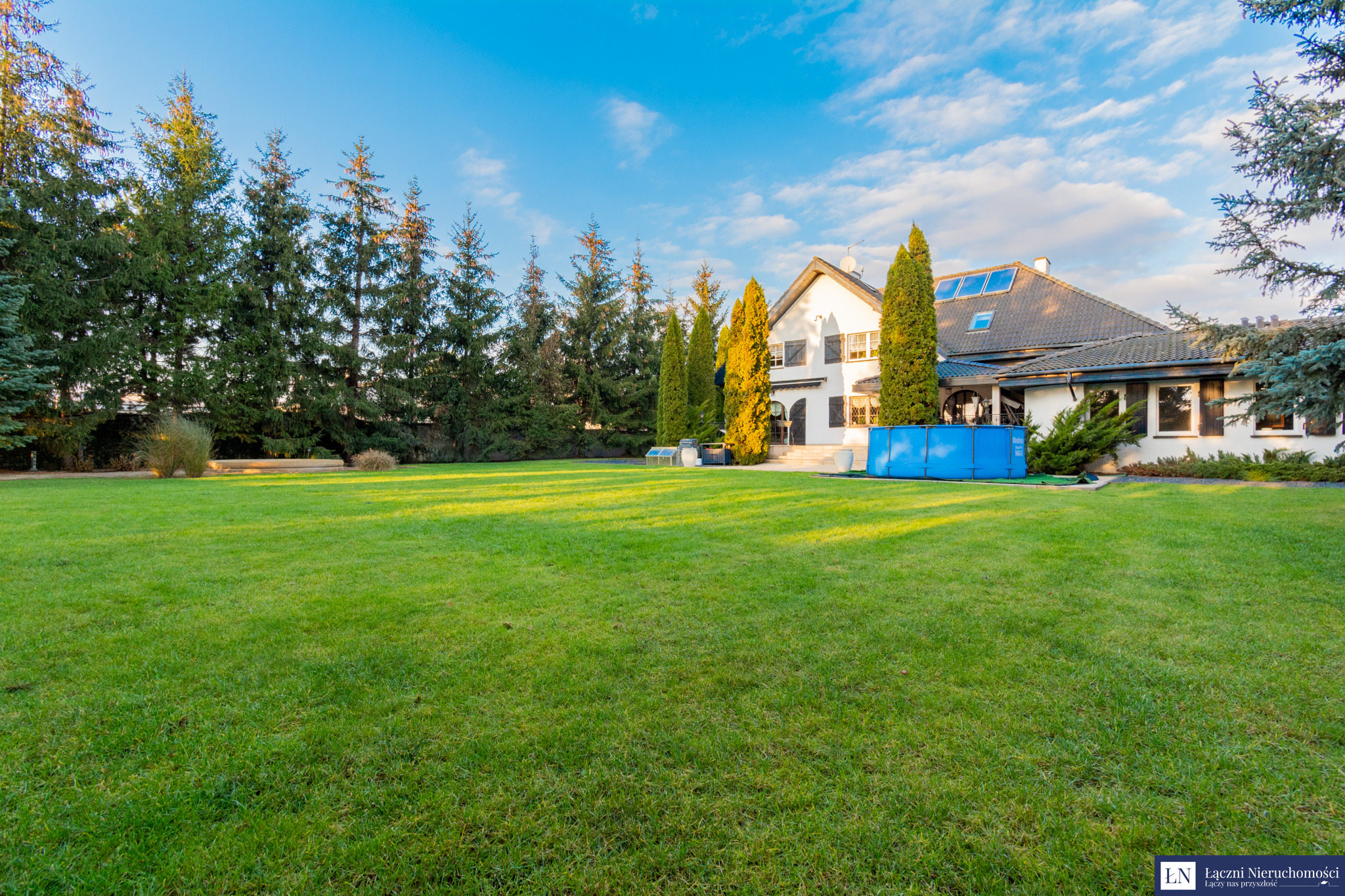 Duży, zadbany trawnik przed dwupiętrowym domem z białymi ścianami, brązowymi okiennicami i szarą dachówką, z widocznym basenem ogrodowym i rzędem wysokich drzew iglastych w tle.