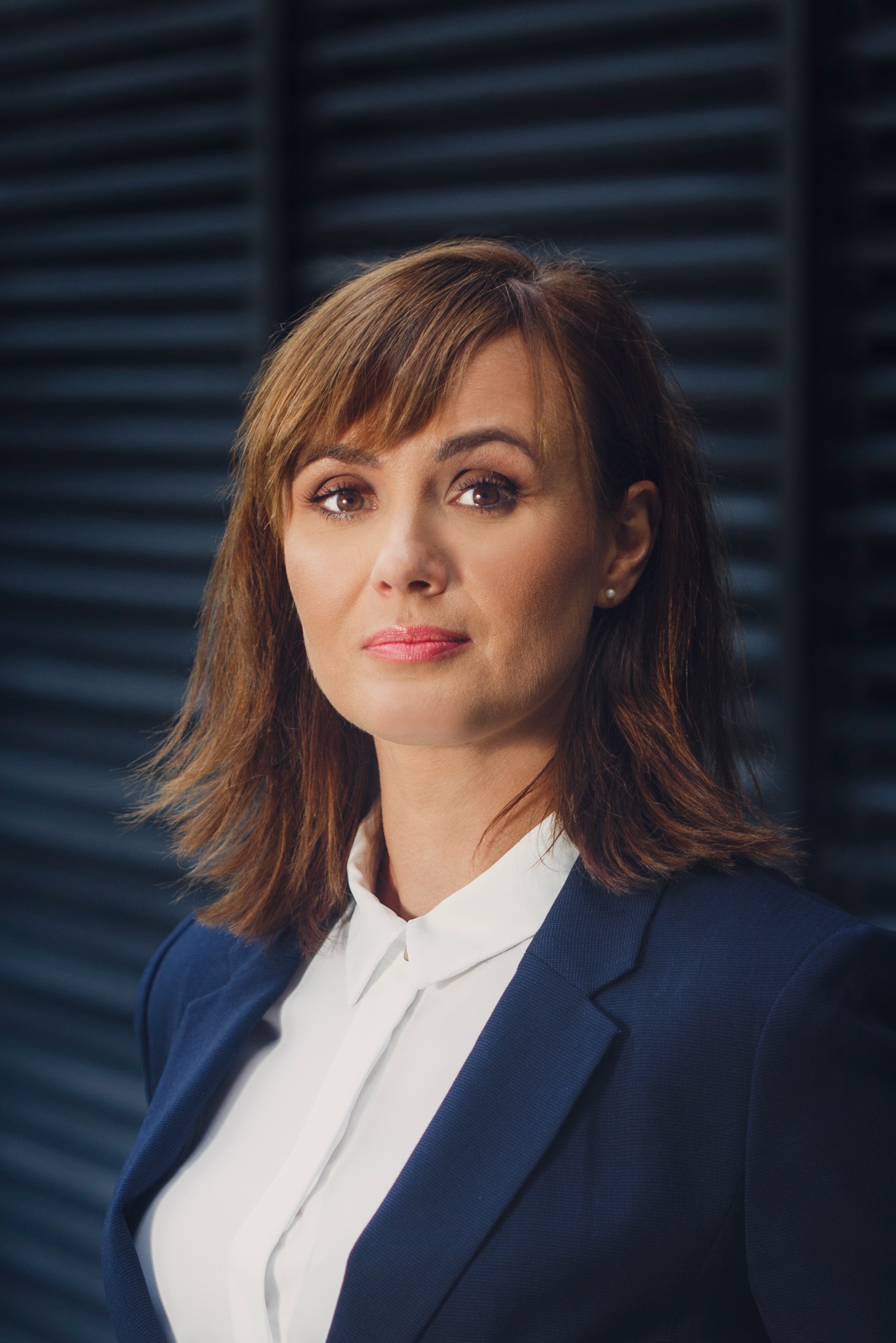 Portret kobiety w granatowym żakiecie i białej koszuli, z delikatnym makijażem i perłową kolczykiem, na tle ciemnej, prążkowanej ściany.