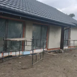 Dom w trakcie ocieplania elewacji, widoczne rusztowania, styropian i częściowo otynkowana ściana w kolorze beżowym, okna zabezpieczone folią.