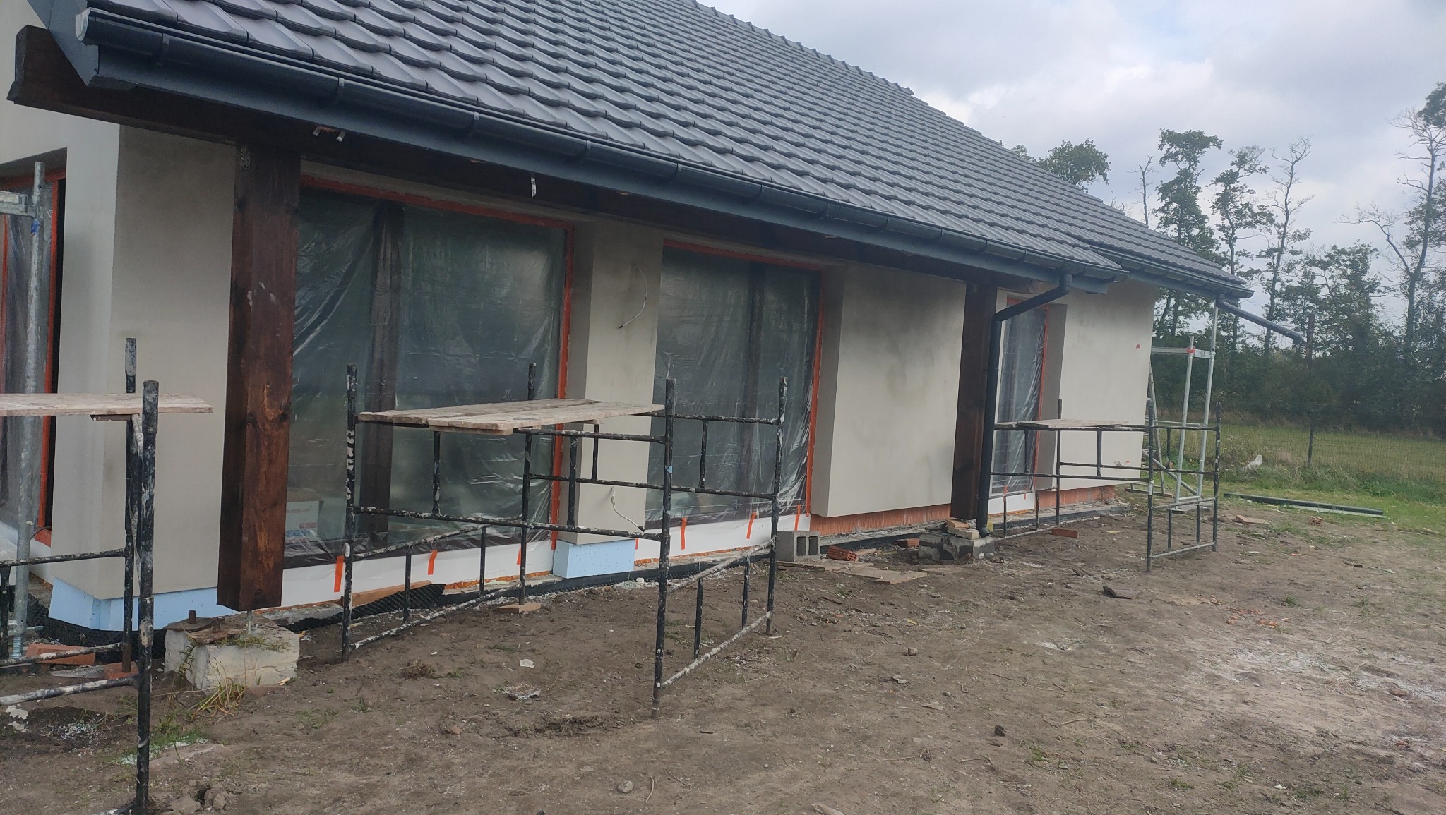 Dom w trakcie ocieplania elewacji, widoczne rusztowania, styropian i częściowo otynkowana ściana w kolorze beżowym, okna zabezpieczone folią.