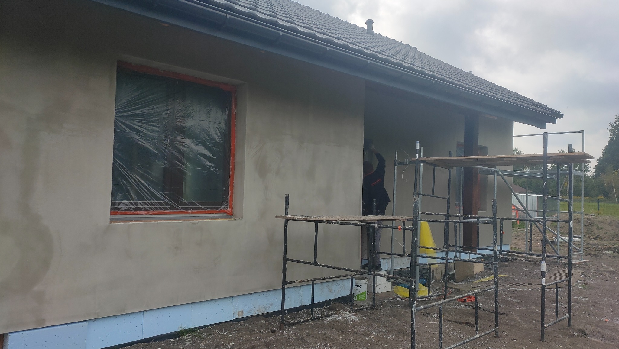 Świeżo otynkowana elewacja domu jednorodzinnego, okno zabezpieczone folią, widoczne rusztowanie i pracownik w trakcie pracy.