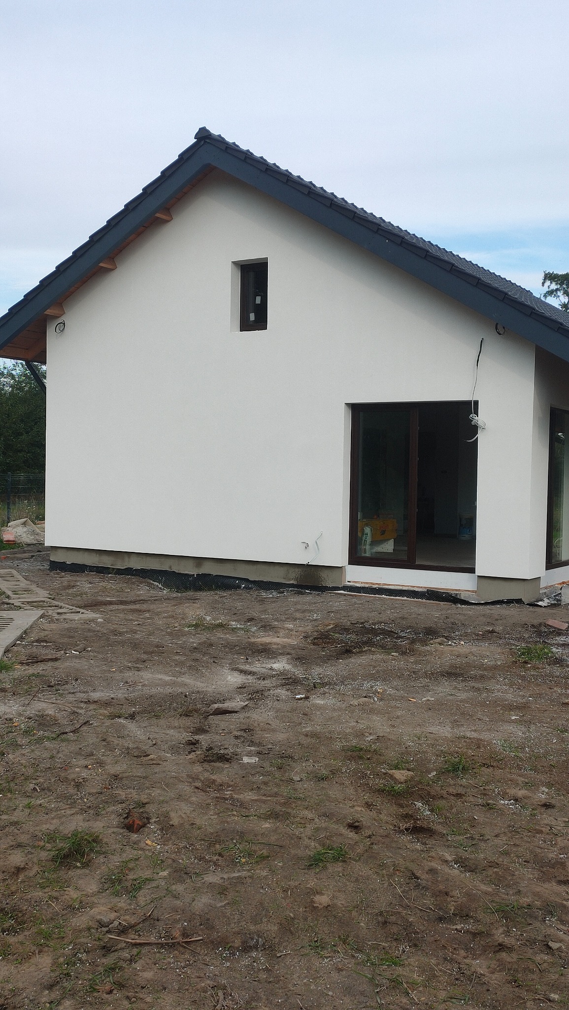 Świeżo otynkowany, biały dom jednorodzinny z ciemną dachówką, widoczne przygotowania terenu wokół budynku.