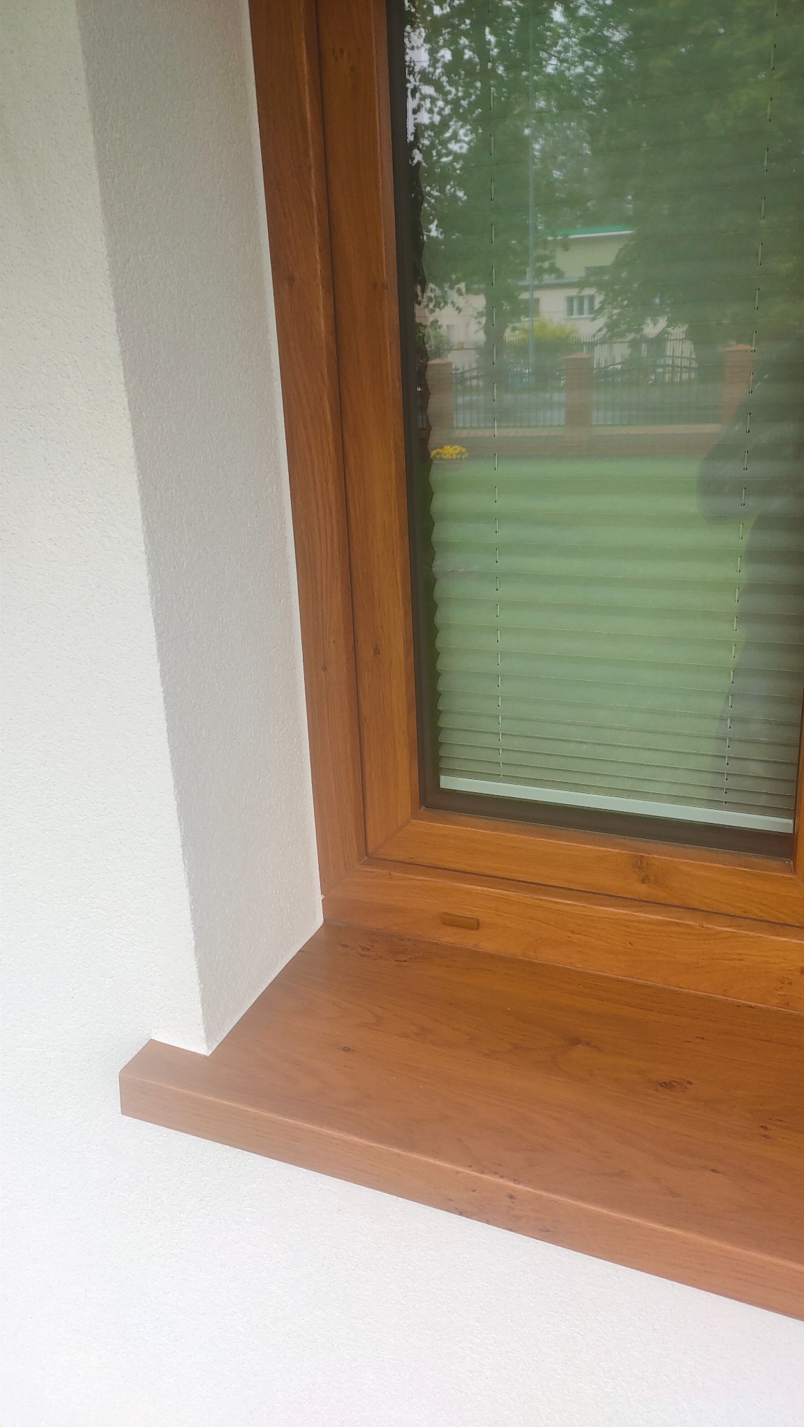 Fragment elewacji z oknem o drewnianej ramie, widoczne rolety plisowane i fragment budynku w tle.