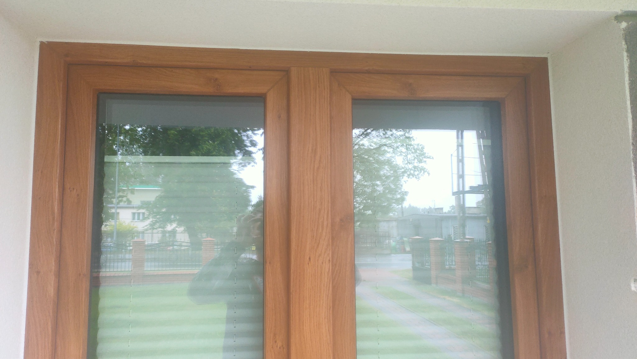 Dwuskrzydłowe okno PCV w kolorze drewna, osadzone w białej elewacji, z widocznym odbiciem fotografa i otoczenia.