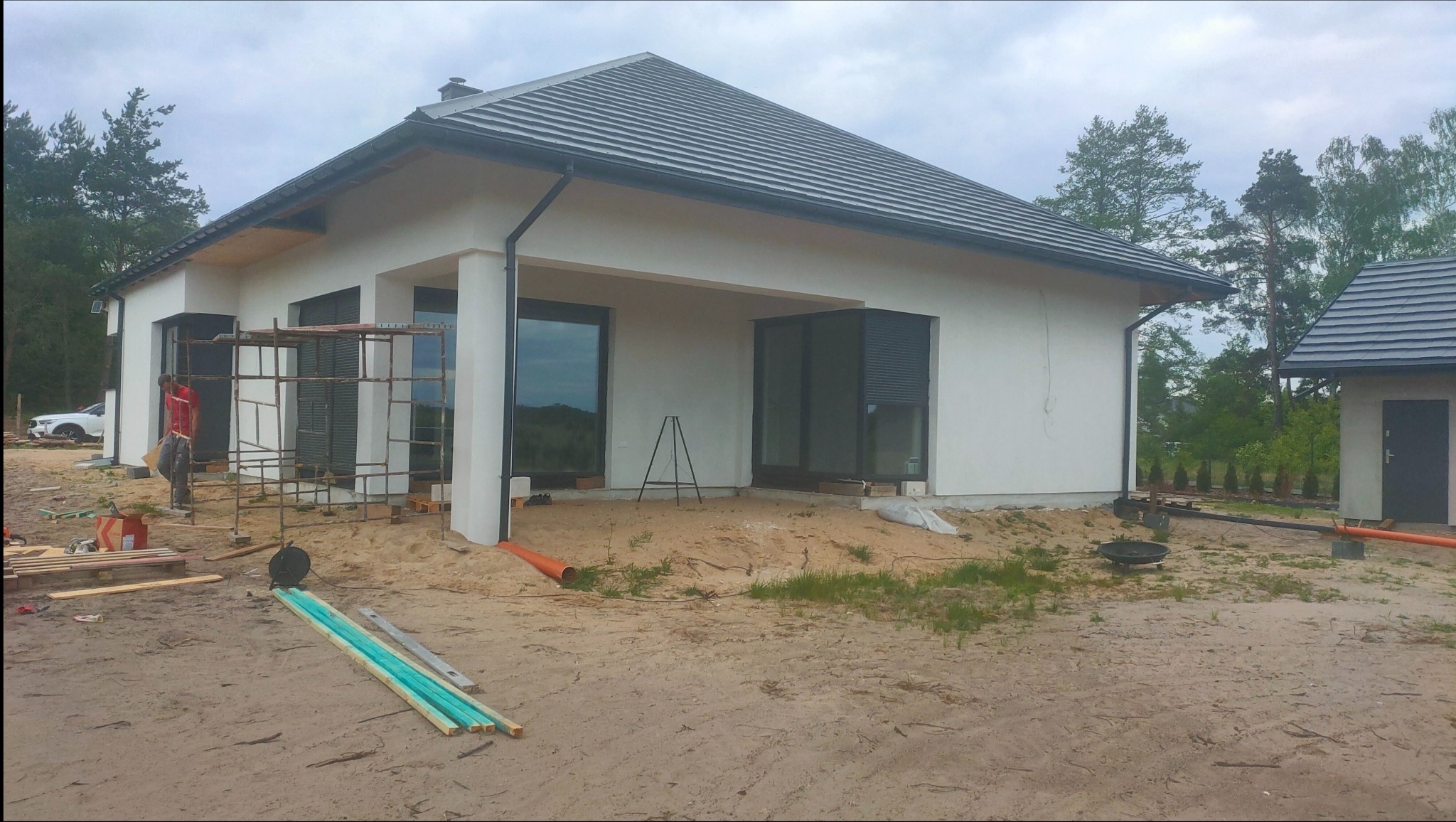 Dom jednorodzinny w trakcie prac elewacyjnych, widoczne rusztowanie, materiały budowlane i pracownik w czerwonej koszulce, teren wokół domu piaszczysty.