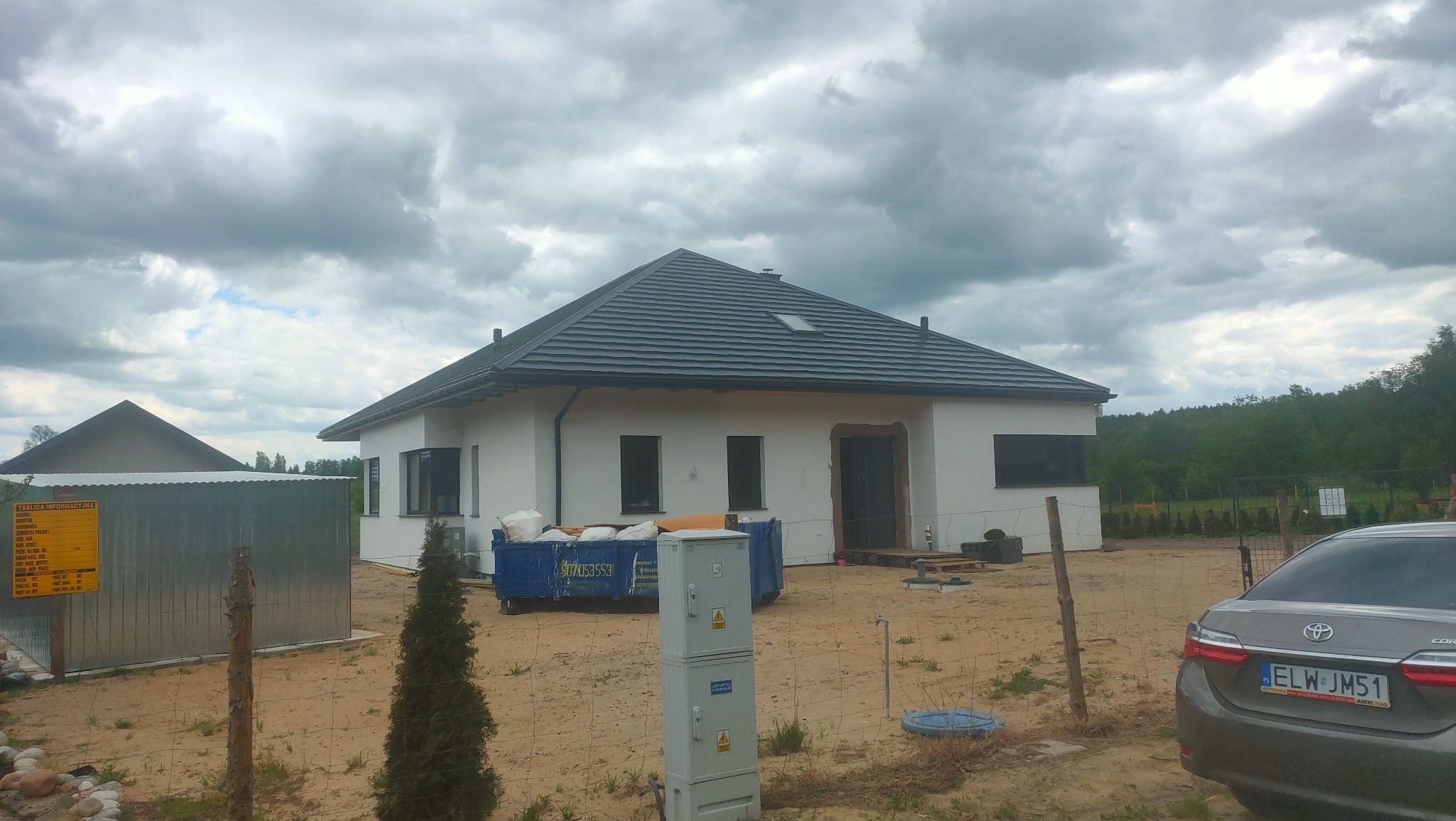 Nowoczesny dom jednorodzinny z białą elewacją i grafitowym dachem, w trakcie prac wykończeniowych, widoczny kontener na gruz i samochód osobowy na pierwszym planie.