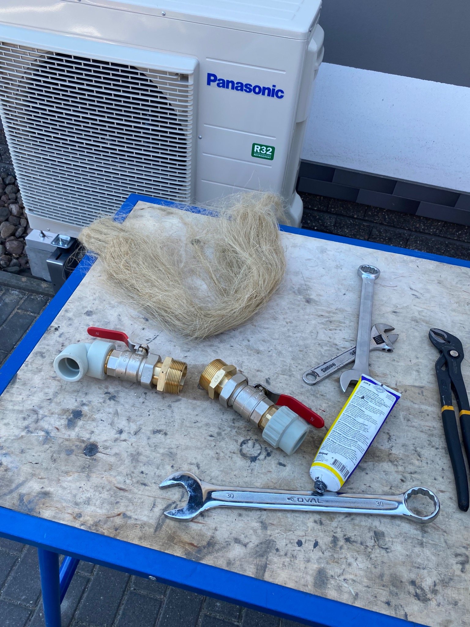 Przygotowanie do montażu pompy ciepła Panasonic: widoczne złączki hydrauliczne z czerwonymi zaworami, pakuły, pasta uszczelniająca Unipak, klucze płaskie i kombinerki na roboczym stole...