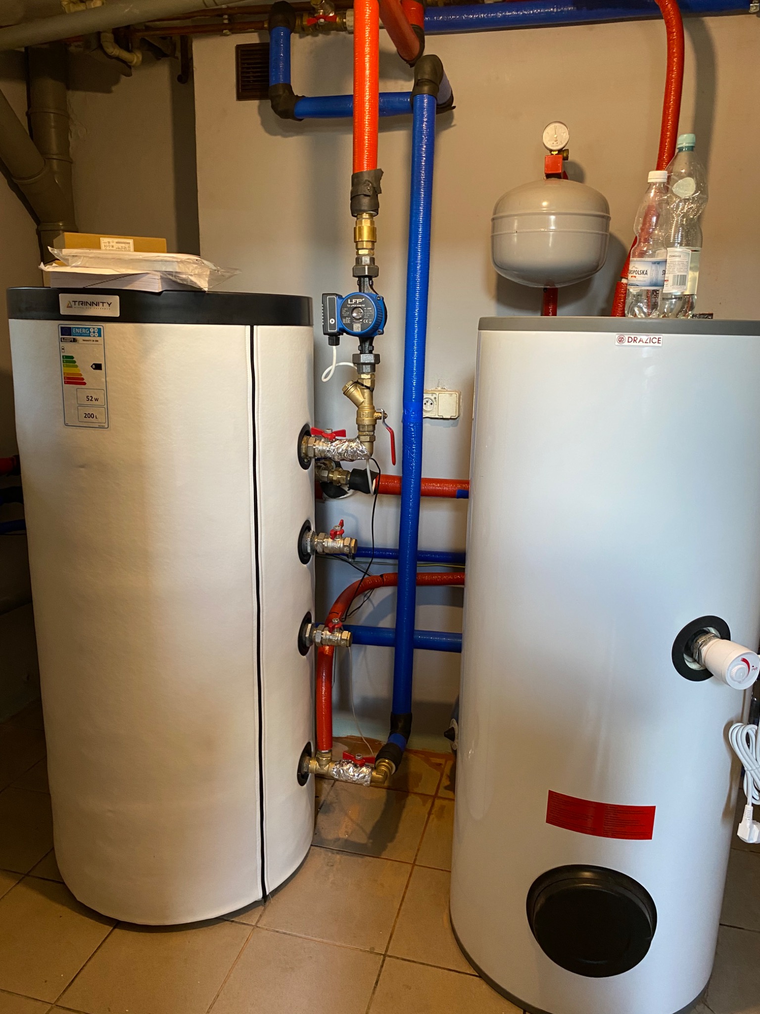 Dwa białe zasobniki ciepłej wody użytkowej (Trinity i Drazice) z podłączeniami hydraulicznymi w piwnicy, widoczne rury w kolorach niebieskim i czerwonym, oraz pompa obiegowa.