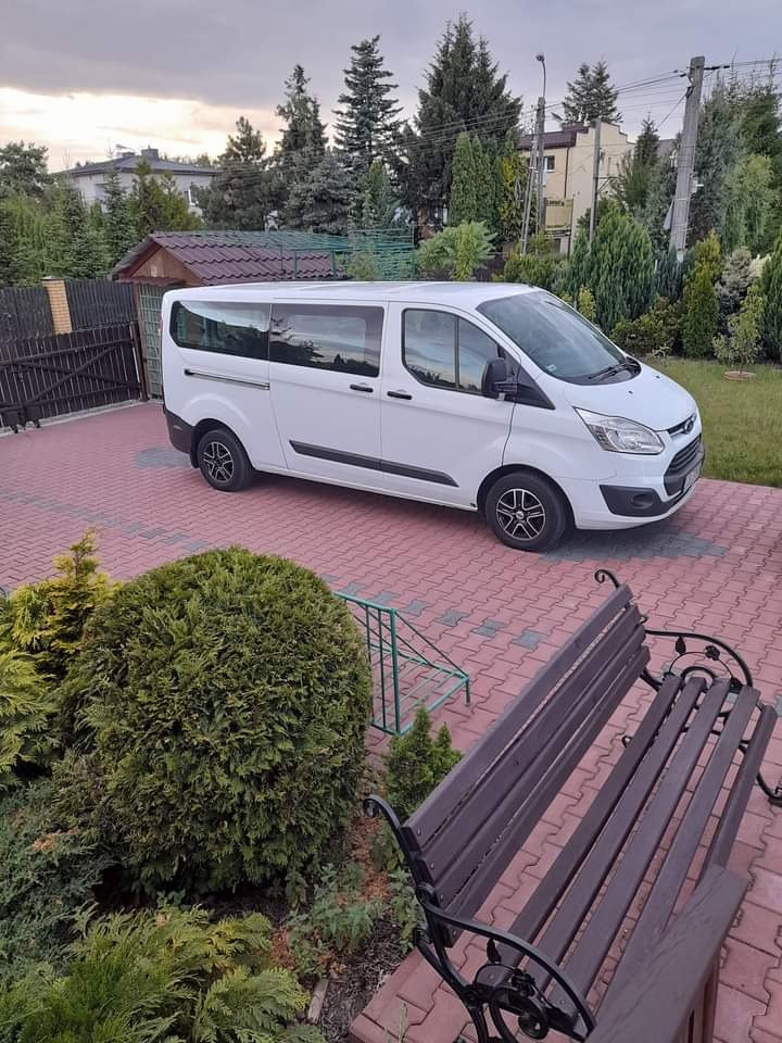 Biały bus Ford Transit na czerwonej kostce brukowej, otoczony zielenią. Widok z boku, lekko z góry. Pochmurne niebo w tle. Charakterystyczne, czarne felgi.