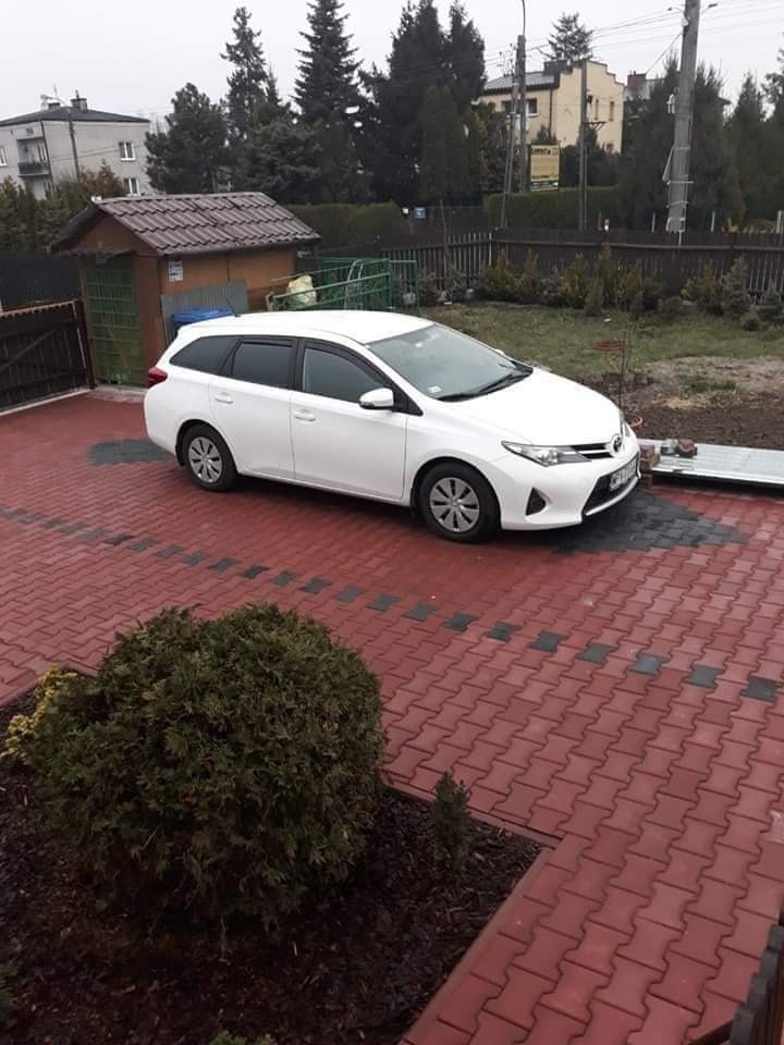 Biała Toyota kombi zaparkowana na czerwonej kostce brukowej przed domem z brązowym dachem i zielonymi elementami. Widok z góry, pochmurny dzień.