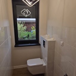 HAUSMAYSTER - Nowoczesna łazienka z podwieszaną toaletą, jasnymi płytkami i oknem z widokiem na zieleń. Uwagę przyciąga designerska lampa sufitowa o geometrycznym kształcie.