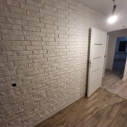 HAUSMAYSTER - Ściana z białej cegły dekoracyjnej i drewnopodobna podłoga, widok na otwarte drzwi do kolejnego pomieszczenia. Widoczny fragment instalacji elektrycznej.