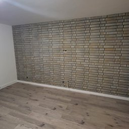 HAUSMAYSTER - Ściana z dekoracyjnej cegły w odcieniach szarości i beżu, z widocznymi gniazdkami elektrycznymi, oraz podłoga z paneli imitujących drewno w jasnych tonacjach.