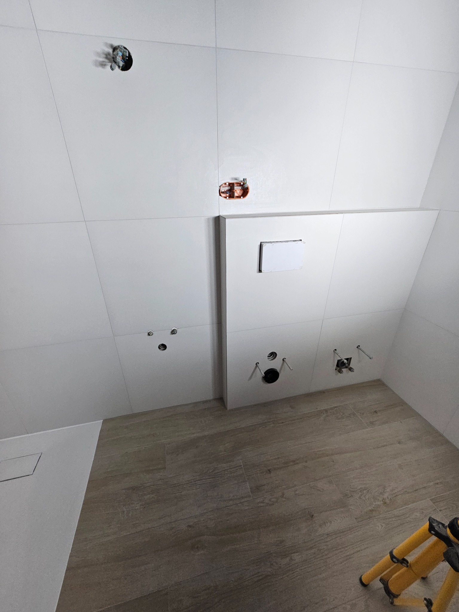 Białe płytki ścienne i podłoga imitująca drewno w łazience w trakcie wykańczania. Widoczne otwory na armaturę i instalacje elektryczne. Statyw w rogu.