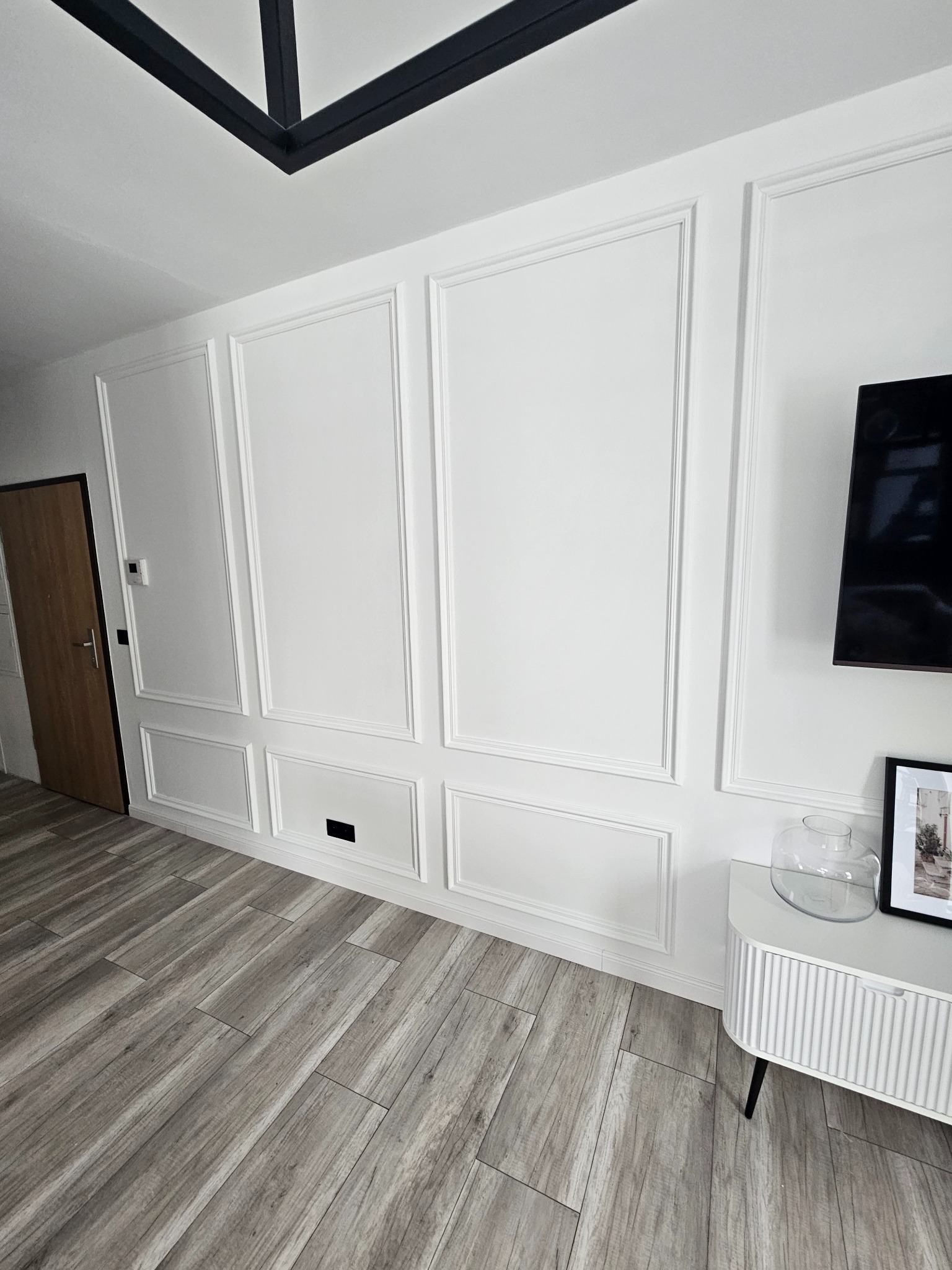 Białe ściany z dekoracyjnymi panelami i listwami przypodłogowymi w nowoczesnym salonie. Podłoga wyłożona panelami imitującymi drewno. Minimalistyczny design.